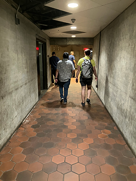 A picture of a pathway in the metro station from the elevator. There are five men in the background of the picture, two of which are walking further away. There are concrete walls on both sides of the picture. A picture of a pathway in the metro station from the elevator. There are five men in the background of the picture, two of which are walking further away. There are concrete walls on both sides of the picture.